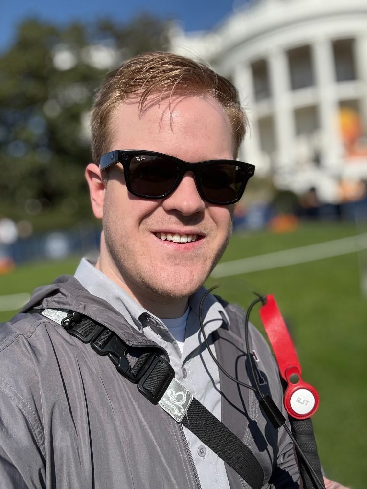 A close-up selfie of James Rath wearing sunglasses and smiling. The White House is visible in the background, along with some Halloween decorations. James wears a gray jacket, and his white cane is visible on his shoulder.