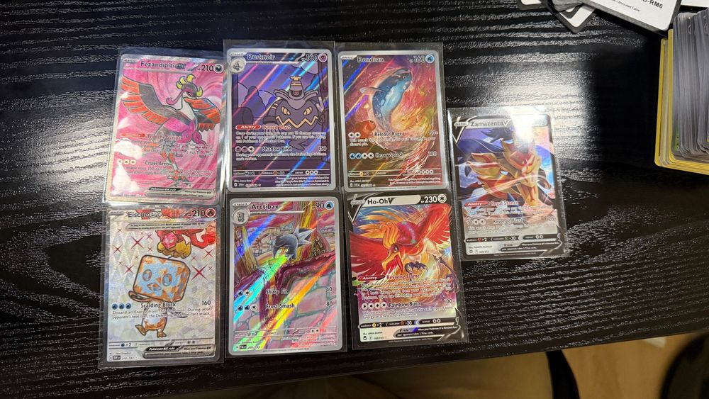 "Six Pokémon TCG cards displayed in a grid on a dark wooden surface. Cards include Fezandipiti ex, Dusknoir, Dondozo, Eiscue ex, Arctibax, Ho-Oh V, and Zamazenta V, each encased in protective sleeves. The cards feature a mix of vivid holographic artwork and unique abilities, highlighting the variety and rarity of the collection."