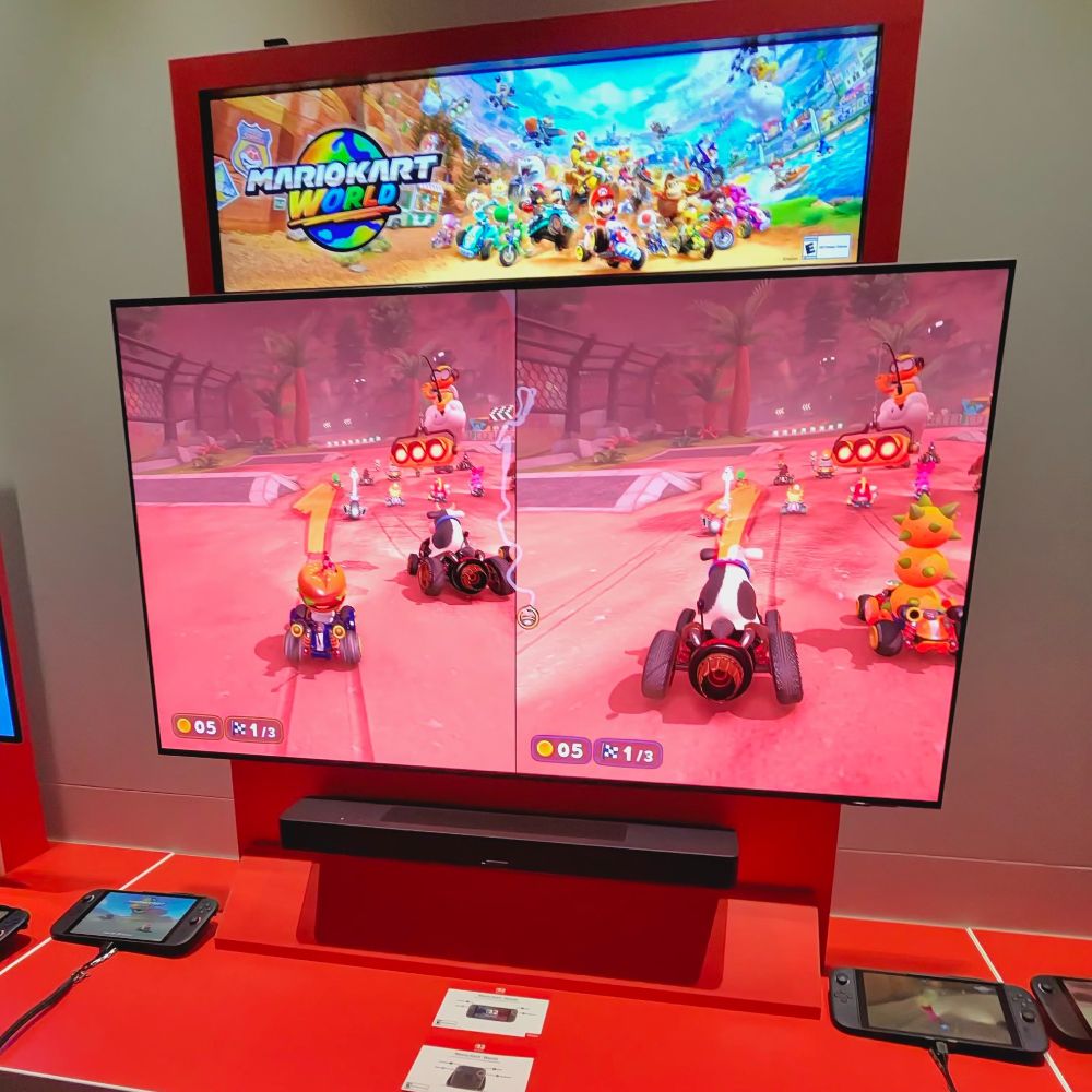 A split-screen view of Mario Kart World Championship gameplay on a large display. The race is starting on a vibrant, pink-tinted track. The station features connected Nintendo Switch 2 consoles and controllers for hands-on play.