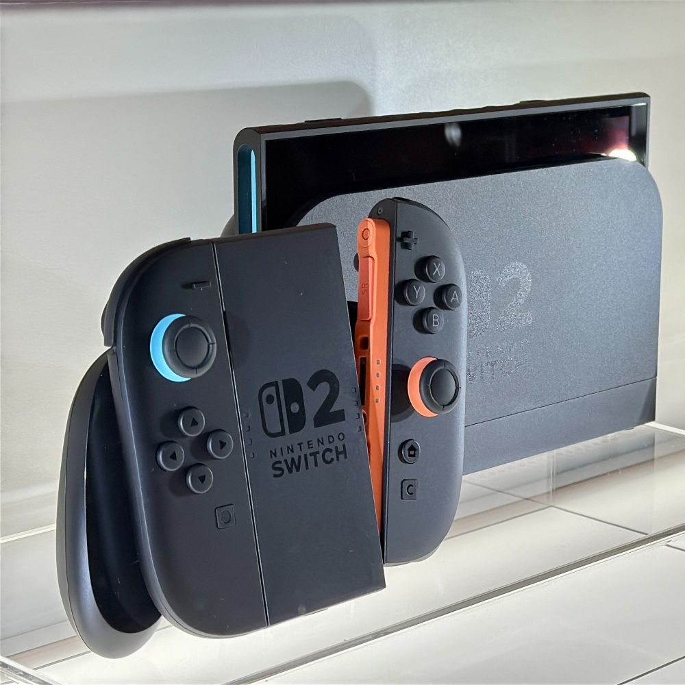 A close-up of the Nintendo Switch 2 console and Joy-Con controllers on display in a clear case. The left Joy-Con is matte black with a blue joystick, the right one is matte black with orange accents, and the console dock is labeled “Switch 2.”