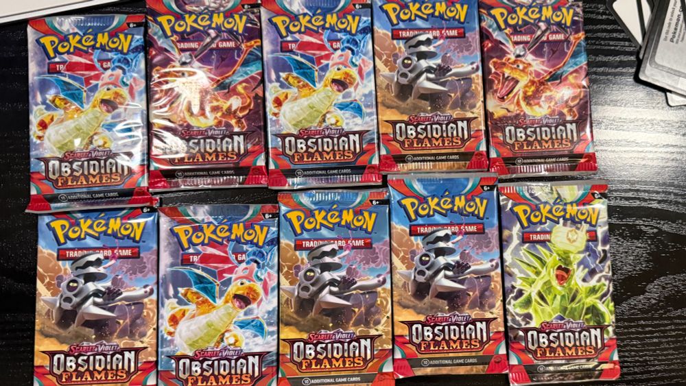 Ten Pokémon TCG booster packs from the Scarlet & Violet: Obsidian Flames series arranged in two rows of five on a dark wooden surface. The packs feature vibrant and dynamic artwork, including Charizard, Tyranitar, and other prominent Pokémon, with fiery and striking designs representing the set.