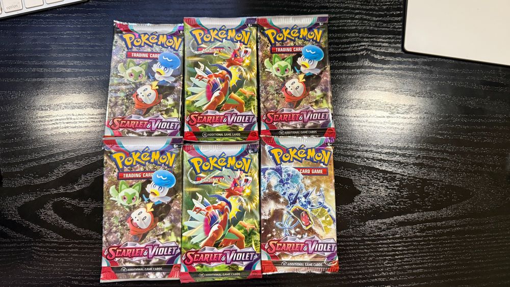 Six Pokémon TCG booster packs from the Scarlet & Violet series are neatly arranged in two rows of three on a dark wooden desk. Each pack features vibrant art of characters and Pokémon from the series, including the starter Pokémon and legendary creatures.