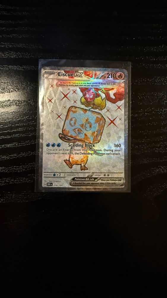 Close-up of an Eiscue ex Pokémon TCG card in a protective sleeve on a dark wooden surface. The card features the playful Ice Pokémon with a unique ice-block head design, surrounded by a holographic background of sparkling stars. The card details its ability and the attack 'Scalding Block,' which deals 160 damage and prevents the opponent's Pokémon from attacking during their next turn.