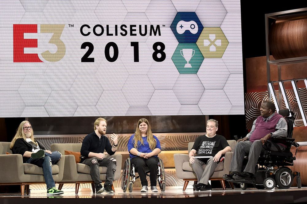 Five panelists sit on stage during the E3 Coliseum 2018 event, with a large screen behind them displaying the E3 logo and gaming icons. James Rath is second from the left, holding a microphone mid-sentence. Other panelists include individuals using wheelchairs and mobility devices, highlighting a discussion on accessibility in games.