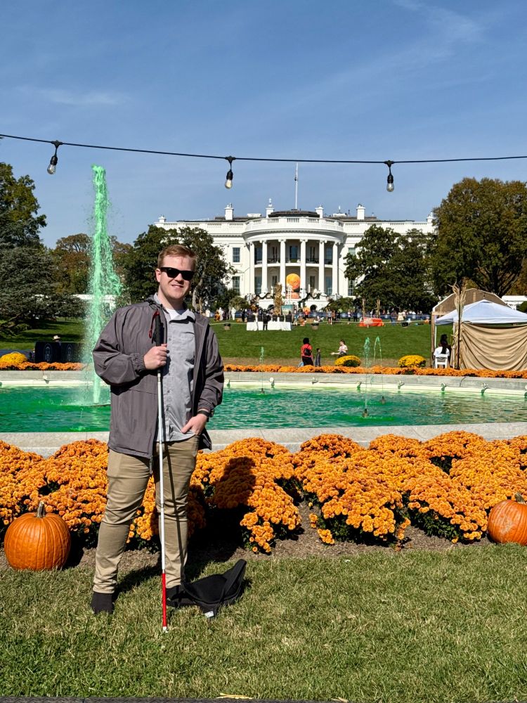 James poses in front of a fountain dyed green, with orange flowers and pumpkins on the grass nearby. The White House is in the background, decorated for Halloween. James is holding a white cane and dressed in a gray jacket and khaki pants, smiling at the camera.

