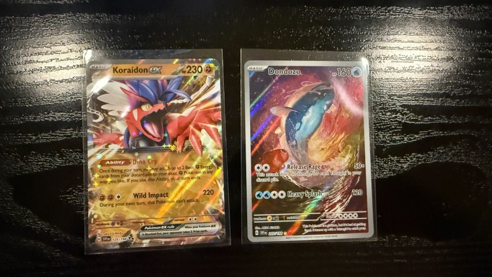 Two Pokémon TCG cards displayed in protective sleeves on a dark wooden surface. On the left is Koraidon ex, featuring the vibrant and dynamic artwork of the ancient legendary Pokémon with a gold foil background. On the right is Dondozo, showcasing the massive aquatic Pokémon swimming through a colorful, ethereal backdrop. A Tatsugiri, sushi-dragon like Pokémon is featured in the left foreground looking up at the Dondozo. Both cards include their unique abilities and moves detailed below their art.