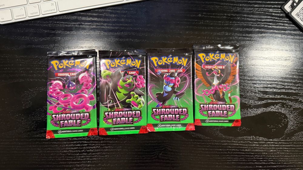 "Four Pokémon TCG booster packs from the Scarlet & Violet: Shrouded Fable series arranged in a single row on a dark wooden surface. Each pack features artwork of various Pokémon, including Houndstone and Fezandipiti, with a mystical and vibrant green and pink theme."