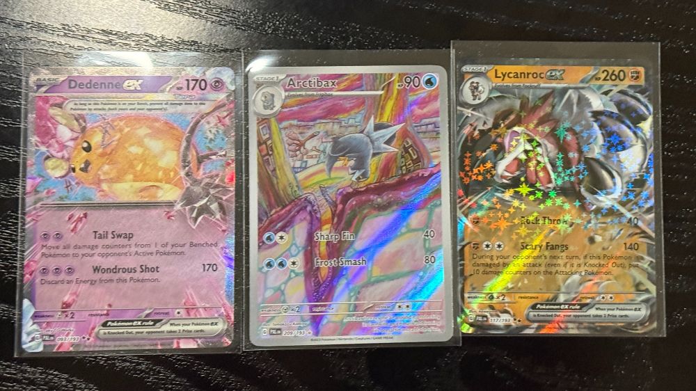 "Close-up of three Pokémon TCG cards displayed in protective sleeves on a dark wooden surface. From left to right: Dedenne ex, featuring the adorable Fairy/Electric Pokémon surrounded by pink blossoms; Arctibax, showcasing the Dragon/Ice Pokémon walking along a vibrant path with an abstract background; and Lycanroc ex, with striking artwork of the Midnight Form Lycanroc, highlighted by a holographic starry effect. Each card includes its abilities and moves detailed below the artwork."