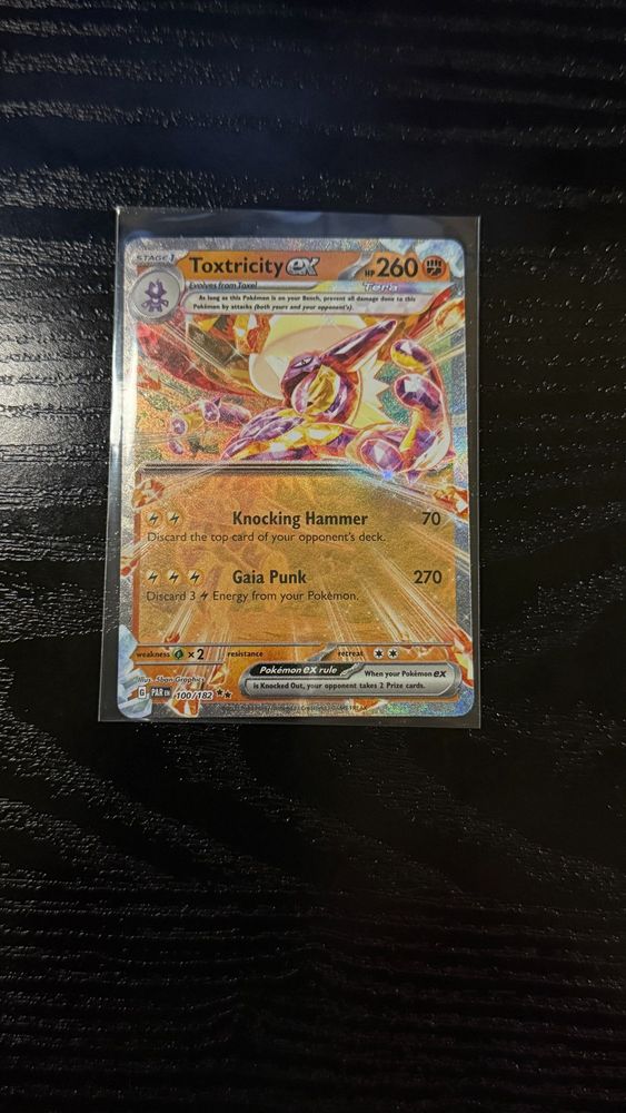 "Close-up of a Toxtricity ex Pokémon TCG card in a protective sleeve on a dark wooden surface. The card features Toxtricity in a dynamic pose surrounded by an electrifying holographic background. The card displays its powerful moves 'Knocking Hammer,' which deals 70 damage and discards the top card of the opponent's deck, and 'Gaia Punk,' which deals 270 damage with a three-Energy discard cost."
