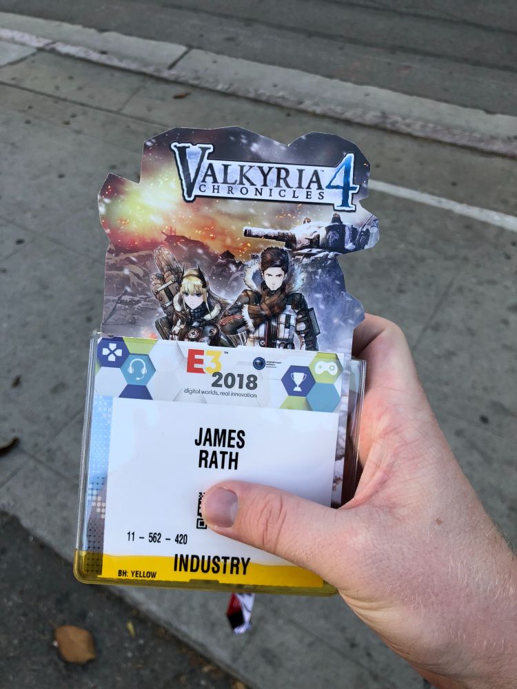 A close-up of James Rath’s hand holding his E3 2018 industry badge, topped with a promotional cardboard cutout for Valkyria Chronicles 4. The badge features the E3 logo, James’s name, and the “INDUSTRY” access label in yellow, with a sidewalk and curb in the background.