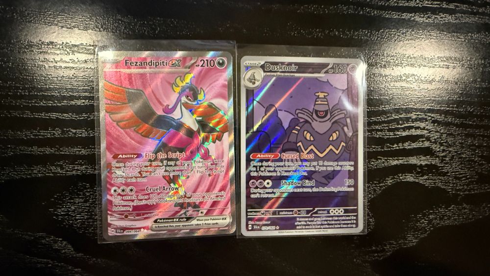 "Two Pokémon TCG cards displayed in protective sleeves on a dark wooden surface. On the left is Fezandipiti ex, featuring the elegant and colorful bird Pokémon with a holographic pink background, and on the right is Dusknoir, showcasing the ghostly Pokémon in a striking purple and blue holographic background with a flat 2D art-style by Pokémon artist, James Turner."