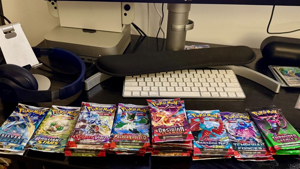 "A desk setup featuring a neat display of Pokémon TCG booster packs arranged in rows. The packs include various sets such as Silver Tempest, Brilliant Stars, Scarlet & Violet, Paldea Evolved, Obsidian Flames, Paradox Rift, Temporal Forces, and Shrouded Fable. There are various amounts of each set from 2 to 10 stacked. In the background, there is a computer monitor, a keyboard, and headphones on a desk stand."