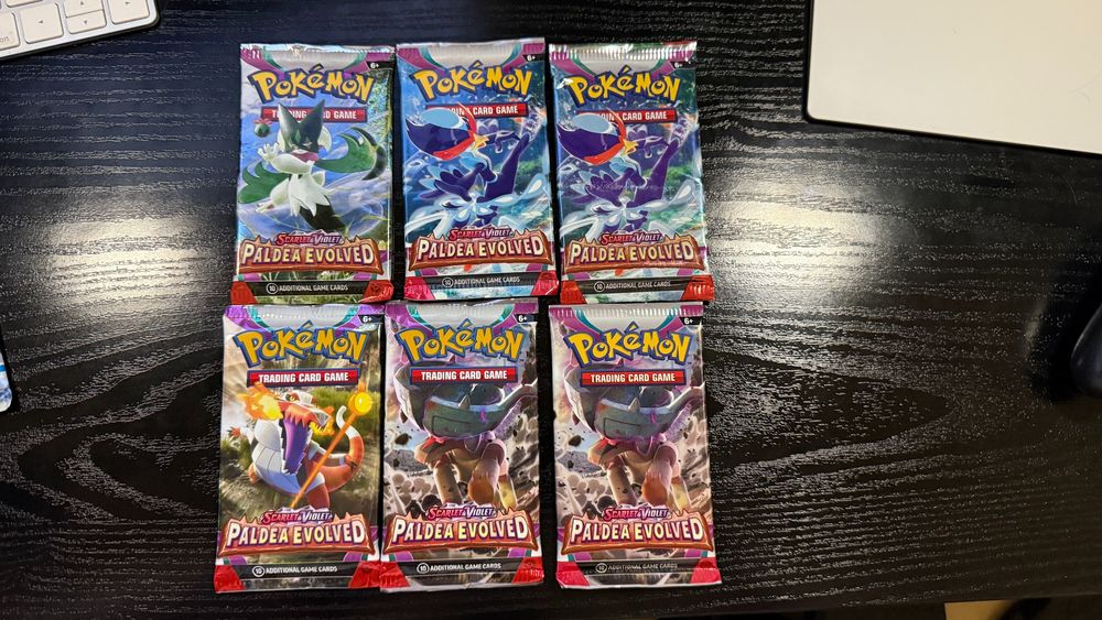 "Six Pokémon TCG booster packs from the Scarlet & Violet: Paldea Evolved series, arranged in two rows of three on a dark wooden desk. The packs feature artwork of various Pokémon from the Paldea region, including the starters and other prominent Pokémon from the set."

