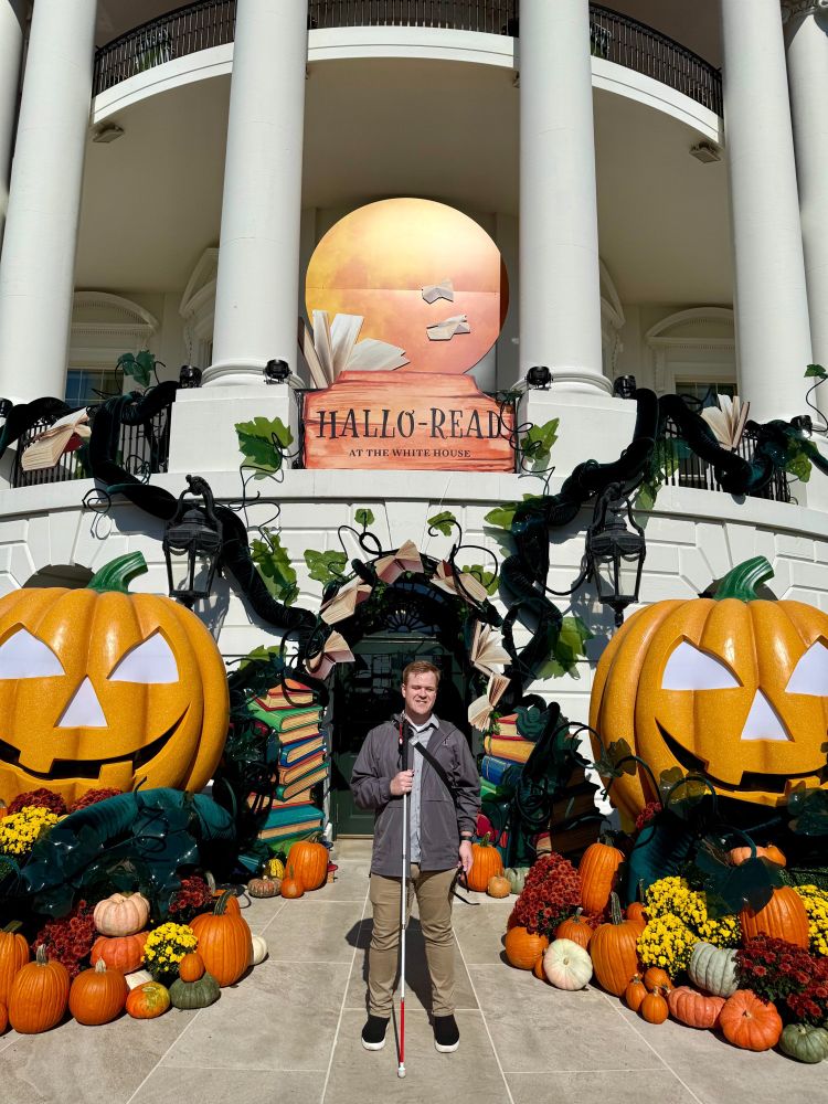 James Rath stands in front of a Halloween-themed display at the White House. The backdrop includes large orange jack-o'-lanterns, stacked books, and a circular sign with the words "Hallo-Read at the White House." James is smiling and holding a white cane, surrounded by pumpkins and autumnal decor.
