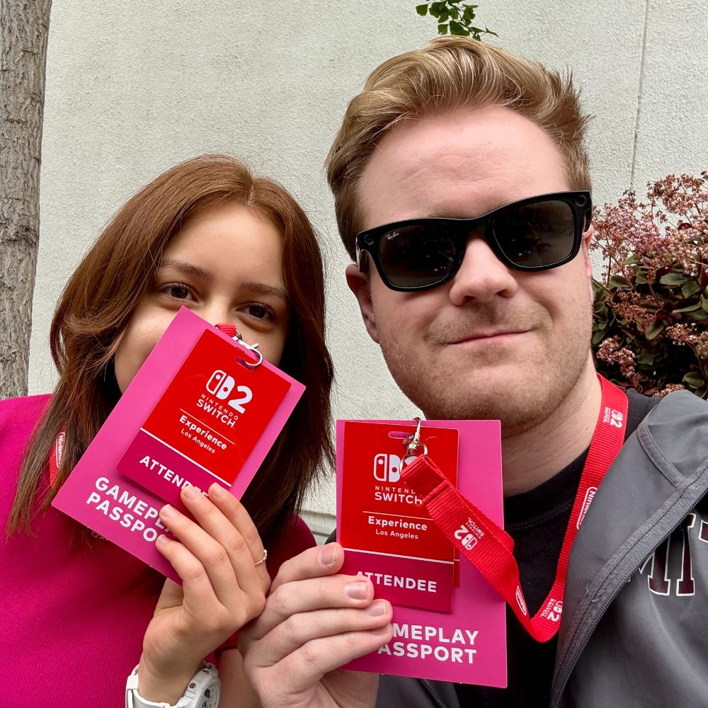 A young man and woman smile while holding up Nintendo Switch 2 "Gameplay Passport" badges in front of them. The man wears sunglasses and a red event lanyard, while the woman wears a pink shirt and white smartwatch. They are attending the Nintendo Switch 2 Experience event in Los Angeles.