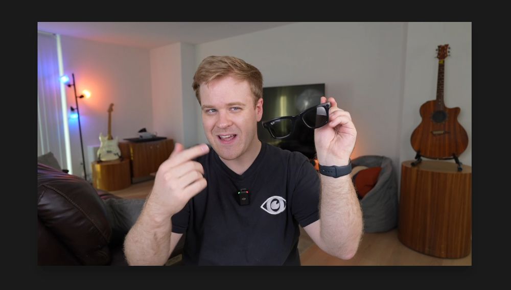 A screenshot of a pause frame from a video. James Rath holds up a pair of Meta Ray Bans and points to them smiling mid-sentence. He sits an entertainment living room with guitars on display, a large TV behind him and colored smart lights colored blue and orange. 