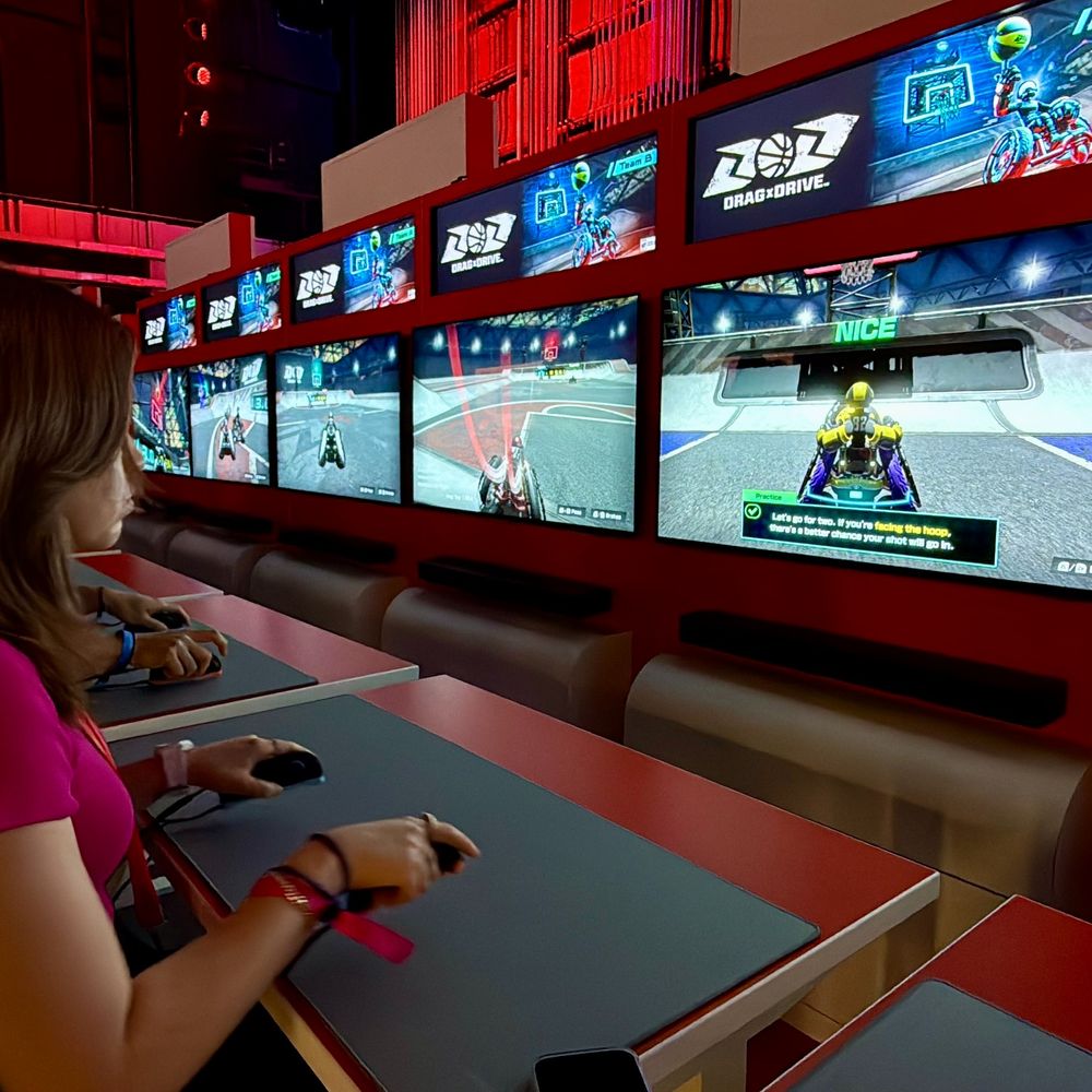 A woman sits at a demo station playing a new Nintendo Switch 2 game titled "Drag & Drive." Multiple large monitors show a stylized basketball-driving hybrid game. The red-lit environment suggests an immersive, arcade-style booth.