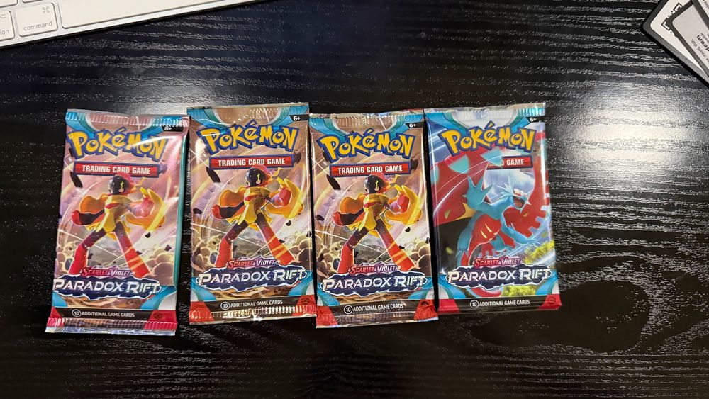 "Four Pokémon TCG booster packs from the Scarlet & Violet: Paradox Rift series arranged in a single row on a dark wooden surface. The packs showcase dynamic artwork of featured Pokémon, including Iron Hands and Mega Sceptile, highlighting the futuristic and prehistoric themes of the set."