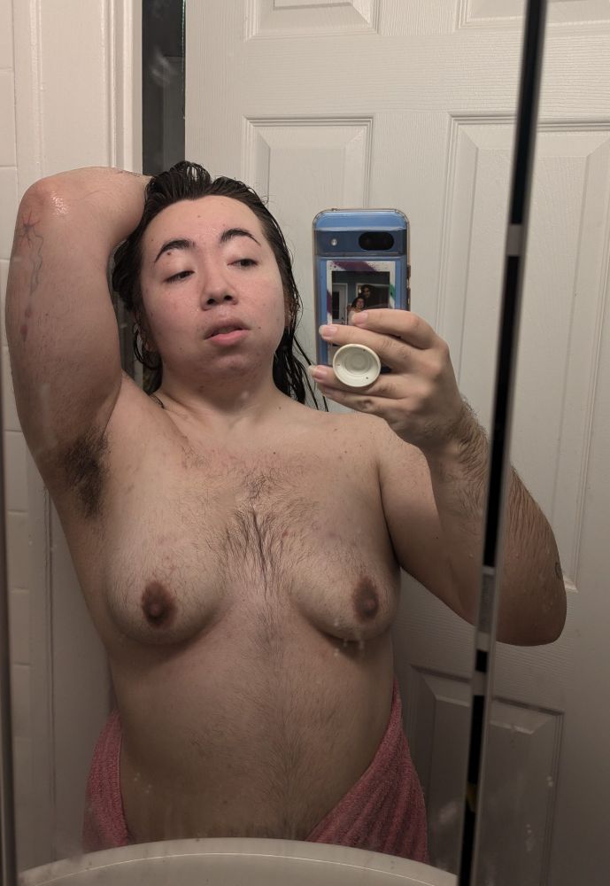 Post shower mirror selfie of Emil, chubby transmasc, arm is lifted to show pit! Pink towel hangs loosely on their hips