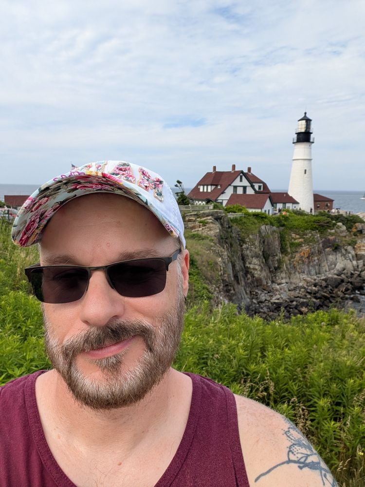 A guy in a tank top, dark tinted glasses and a floral brimmed hat. Oh, and there's a lighthouse in the background.