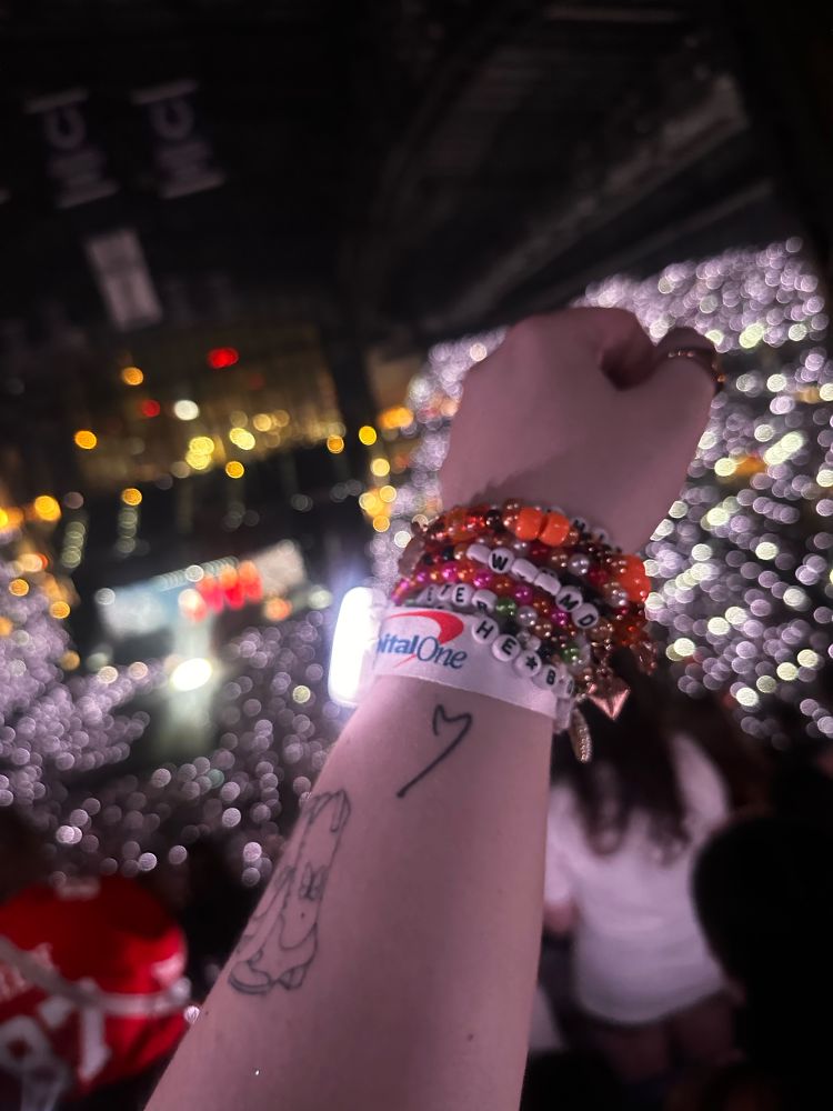 photo from the era’s tour of a tattoo of a heart from a signed TTPD vinyl- there are multiple different colored friendship bracelets & the stadium is lit up white