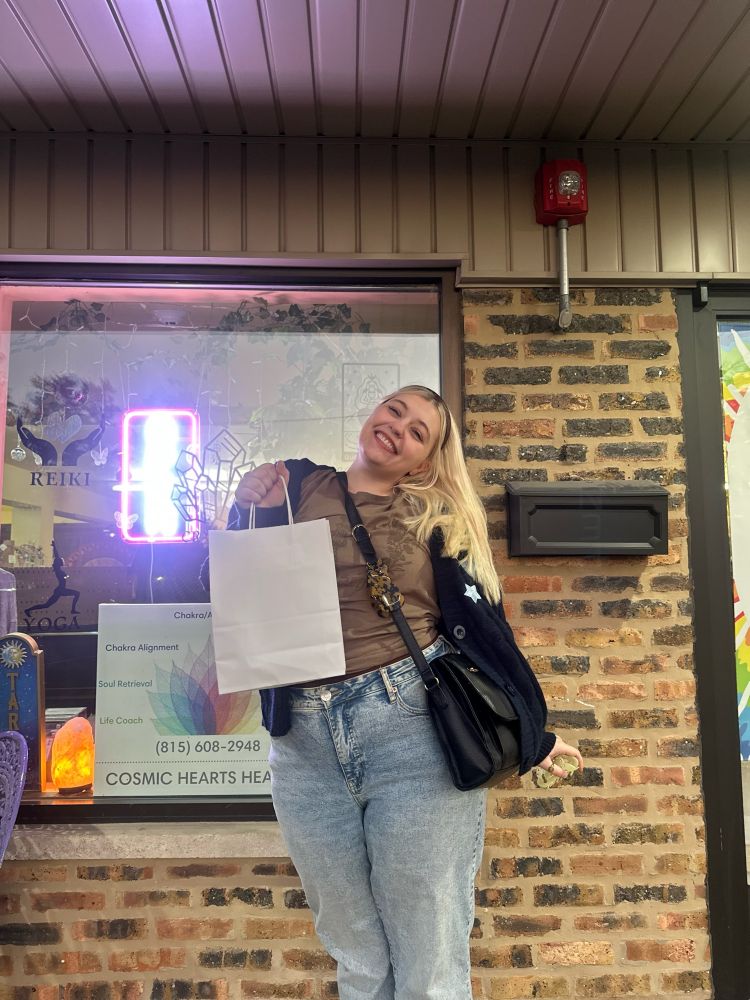 bluesky user scorpioswiftie on her 26th birthday!! she has blonde hair & is wearing a brown “tight rope” taylor swift shirt, a blue midnights taylor swift cardigan, and light blue/acid wash jeans! she is standing in front of a crystal store
