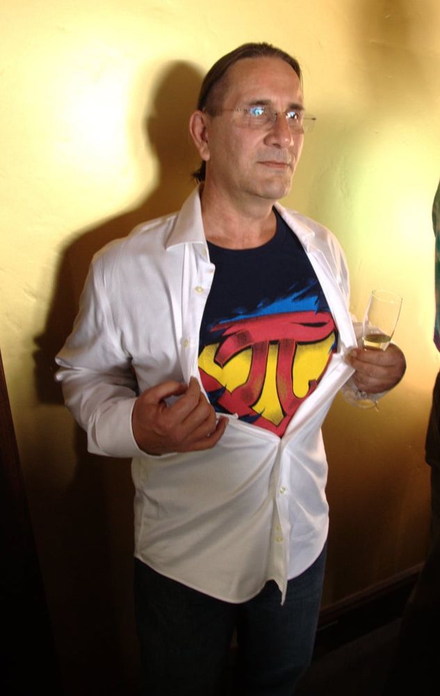 William, wearing a Super Pi shirt under a white dress shirt, standing against a yellow wall.