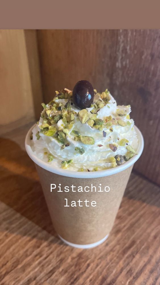 A paper Café cup with a pistachio latte, covered in whipped cream and chopped pistachio nuts,topped with a chocolate covered espresso bean. 