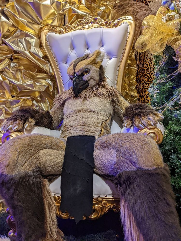 A hippogryph fursuiter wearing nothing but a long loincloth sitting on a chair staring down at you from a dramatic angle 