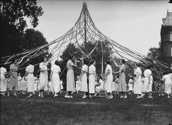 Gray scale glass plate negative of women from the class of 1936 in light colored ankle length dresses intertwining colorful ribbons around a May pole in a field of grass. 
