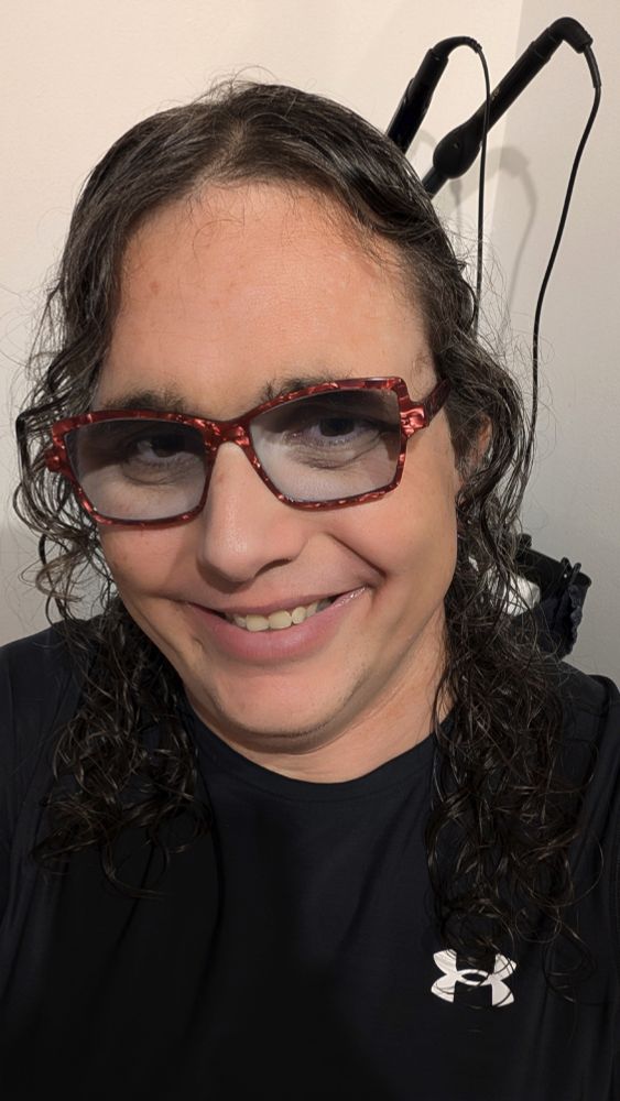 Selfie of Drew with curly hair coming down over her shoulders. She is smiling at the camera, and is wearing red framed glasses, and a black tshirt. 