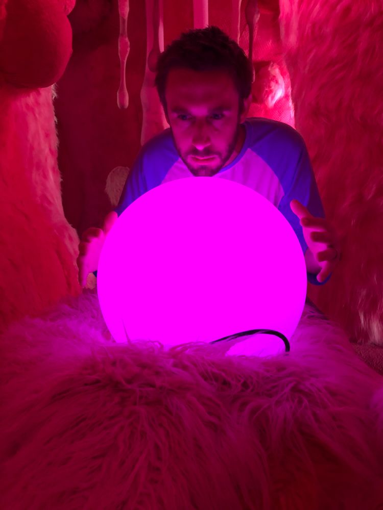 I'm leaning over a glowing pink orb that's as large as my torso with my hands extended over it. I have a blue and white baseball tee and I'm seated in a room with magenta fur on all the walls.