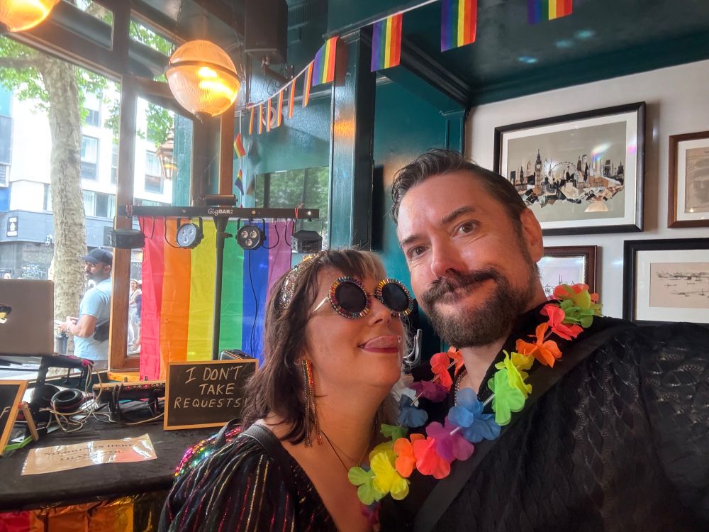 Me wearing a rainbow garland next to a friend in a pub with rainbow flag behind us