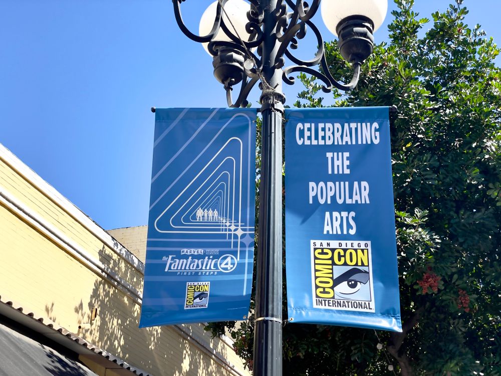 A pair of San Diego Comic-Con banners on a gaslamp-style lamp post. The left one has the Fantastic 4 movie logo in white on a blue background, and the right is the standard “Celebrating the Popular Arts” slogan and Comic-Con logo on matching blue. 