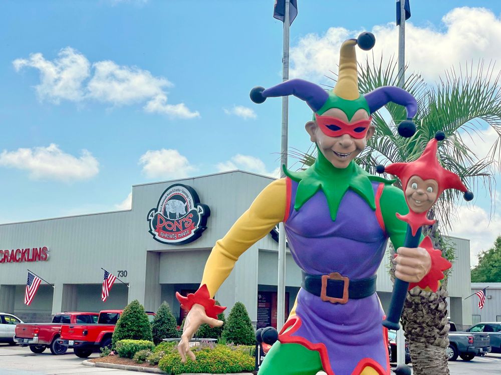 In the foreground is a jester statue in purple, green and gold Mardi Gras colors. In the background is Don’s Specialty Meats.