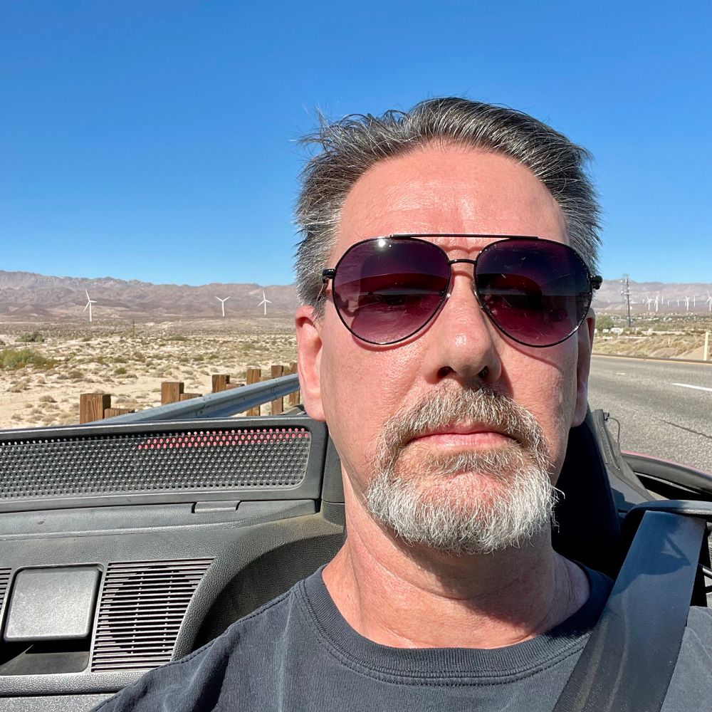 A selfie of middle-aged man with gray hair, gray beard and aviator style sunglasses, pulled over in a convertible, with mountains and a wind farm in the distance behind him. 