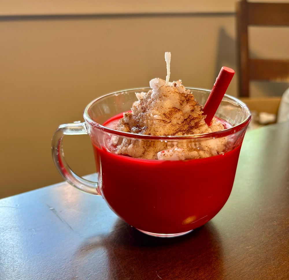A candle that is made to look like a glass mug full of apple cider with whipped cream and cinnamon on top.