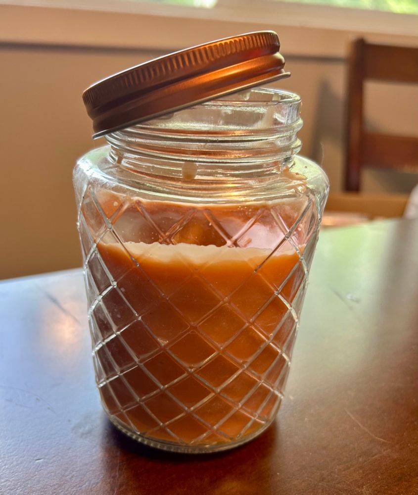 A brown-orange colored candle in a pretty, textured jar with the lid cocked to the side. 
