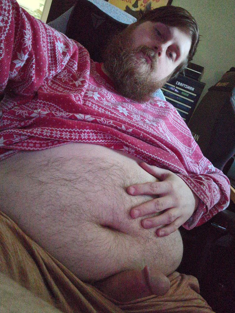 Brown haired bearded bear in a comfy Christmas sweater reclines, his furry round belly exposed. His hand rests on his stomach and his cock hard peeks from his shorts.