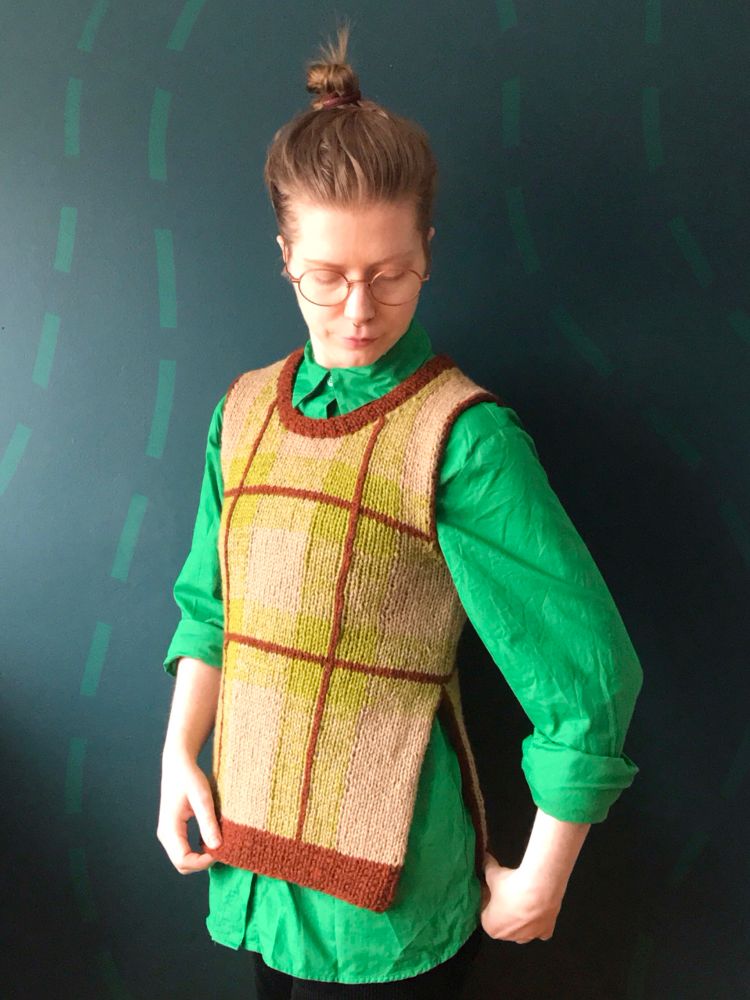 Sara is standing in front of a dark teal wall wearing a bright green shirt and a beige, light green and rust coloured plaid knitted vest. They are looking down and holding the ribbing at the bottom of the vest. 

#knitsky #knitweardesigner 