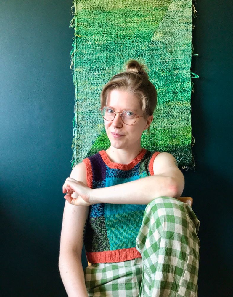 A Caucasian person sits on a chair in front of a green, scrap yarn wall hanging. They have one leg up and rests an elbow on the knee. They are wearing white and green gingham pants, an orange, purple, green and turquoise plaid, knitted vest and are looking into the camera with a smile.