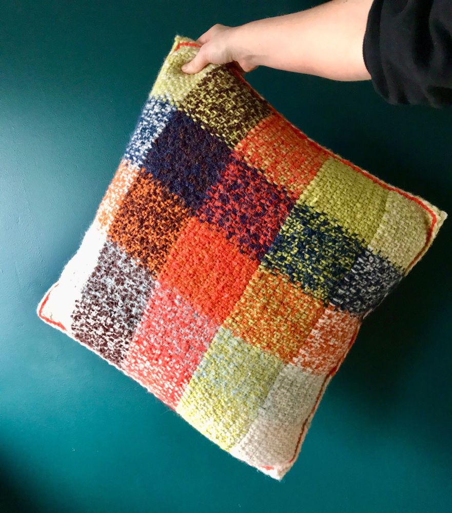 A knitted plaid pillowcase in white, orange, brown, navy, light blue and light green is held up in front of a dark teal wall.