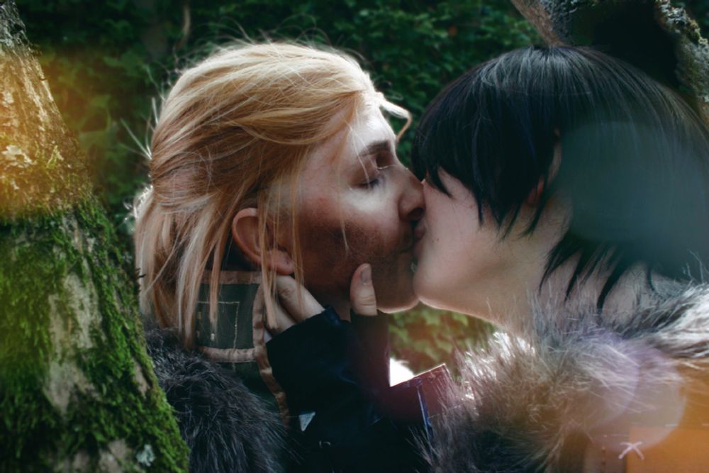 Cosplayers dresses as female Hawke and Anders from Dragon Age 2. They are standing in between some trees. One of Hawke's hands is caressing Anders' jaw as they kiss.