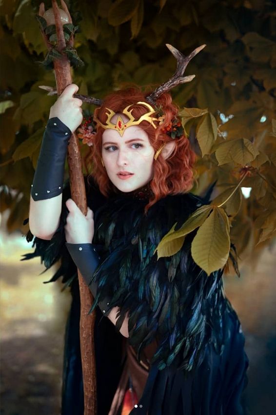 A picture of me in my Keyleth cosplay (Critical Role). I am wearing a black feather cloak over Kiki's brown druid robes and am leaning on a staff. The colours and atmosphere are soft and warm and almost ethereal.