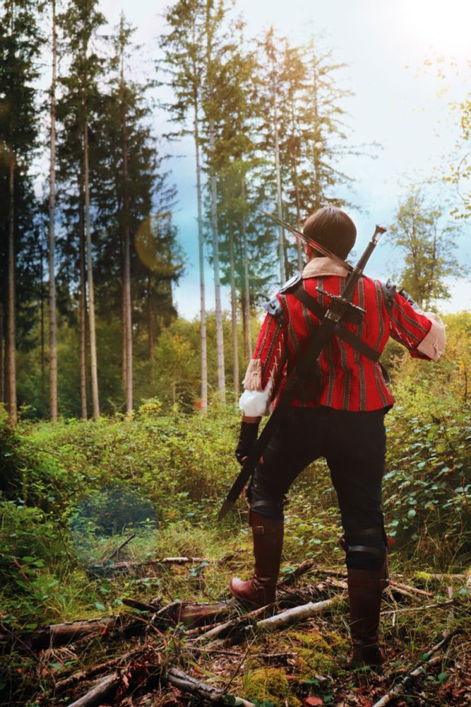 Picture of a person in Eskel (Witcher 3) cosplay. He is pictured from behind, walking away from the camera towards a forest, one sword drawn and resting on his shoulders.