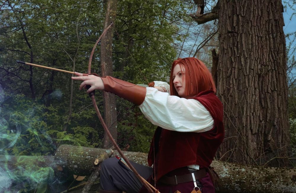 Photo of a white person cosplaying Leliana from Dragon Age Origins. They are wearing a white shirt with billowing sleeves, a dark red velvet doublet and light brown leather bracers. The mood is cool and suspenseful, set in a forest. Leliana is pictured from the hips up, her bow raised and an arrow ready to let loose, her head cocked slightly in concentration.