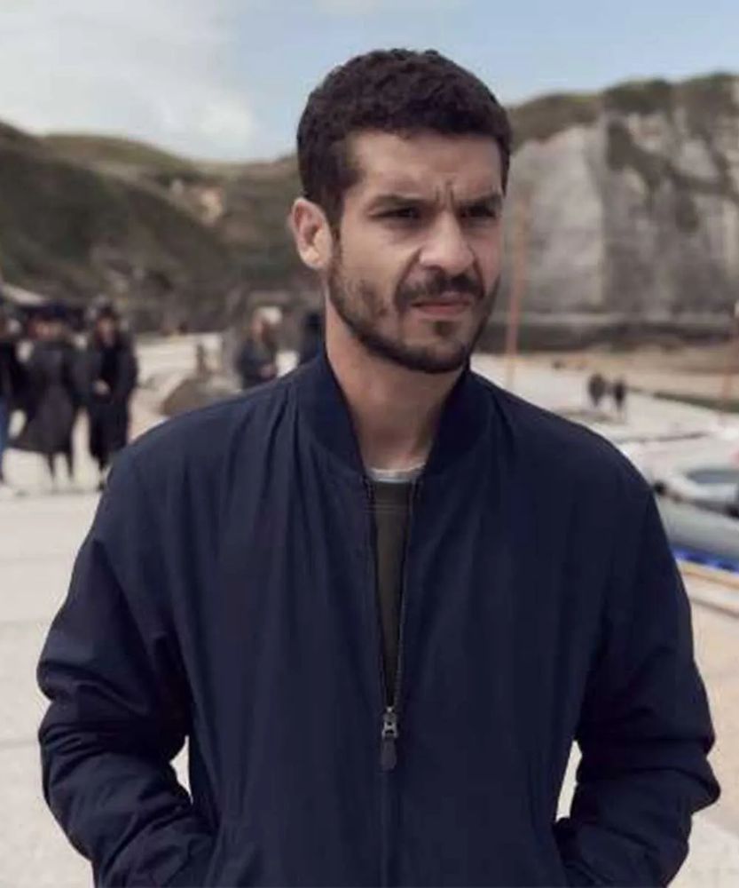 Soufiane Guerrab as Detective Youssef Guédira in Lupin.

A man stands on a beach. He is wearing a blue jacket over an olive shirt. The shirt is barely visible because the zipper is only halfway down. His hair is short and curly and he has a short beard. He is looking off to the distance, with a serious expression on his face.