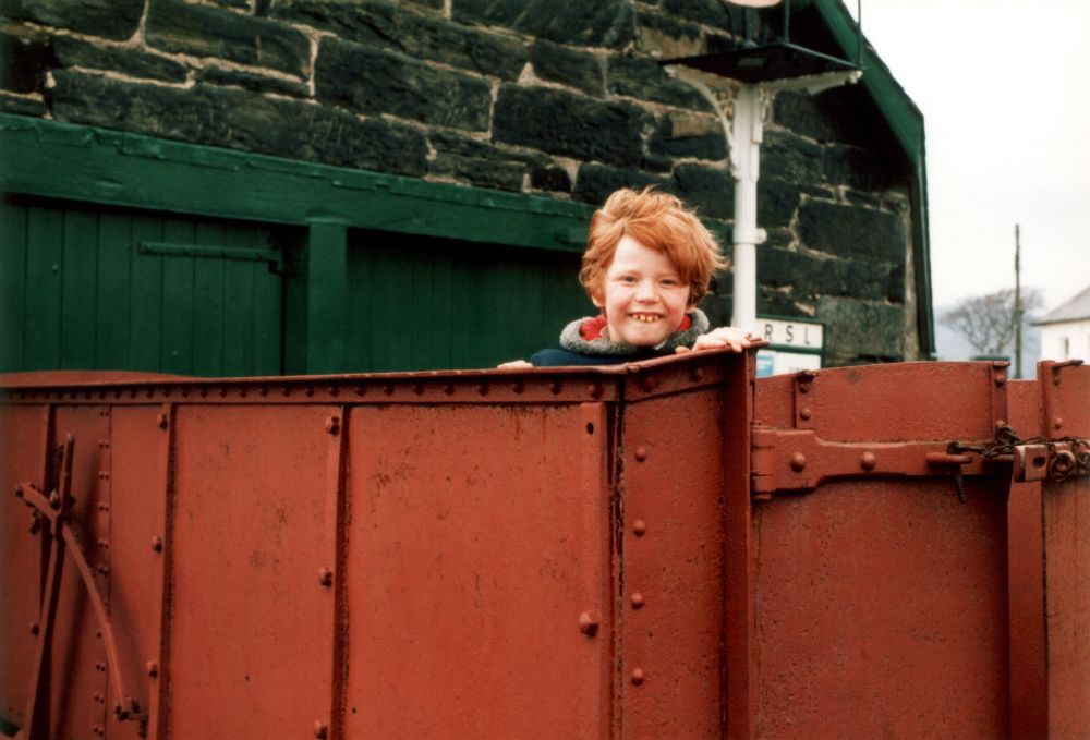 Goofy looking child with a ginger bowl cut, poking his head out of an old railway wagon. 