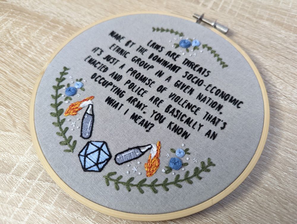 an embroidery hoop with gray fabric with the Laws Are Threats quote from Dimension 20's Fantasy High season embroidery on it with embroidered dice and molotov cocktails