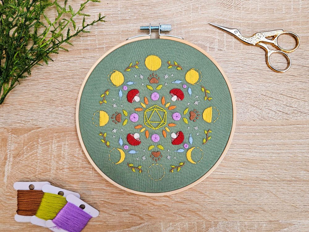 an embroidery hoop with green fabric and colorful druid themed embroidery including the phases of the moon, mushrooms, paw prints, feather, leaves, and flowers